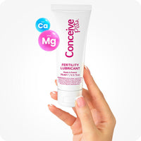 Conceive Plus Fertility Lubricant Tube (FR) held upright between two fingers, showing the 75ml size clearly printed on the front. The white tube features pink text highlighting its sperm-friendly formulation made in France. Ca⁺ and Mg⁺ ion graphics are placed to the left, indicating the key fertility-supporting minerals.