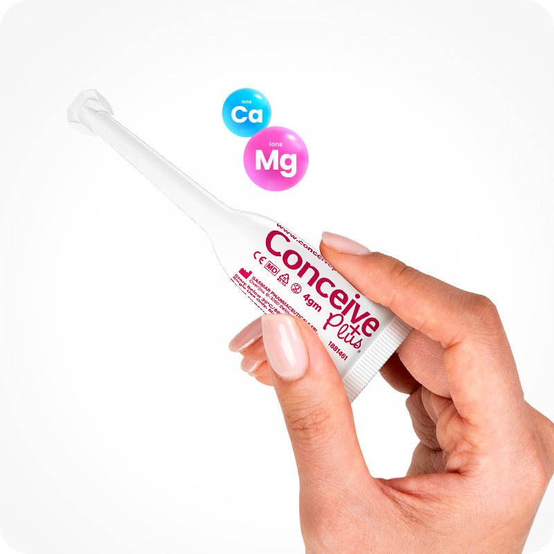 Close-up of a hand holding a Conceive Plus Fertility Lubricant, Pre-Filled Applicators (FR) 4g unit. The single-use applicator is labeled in French and Dutch, featuring calcium (Ca) and magnesium (Mg) ion graphics above. Designed to support natural fertility and sperm viability.